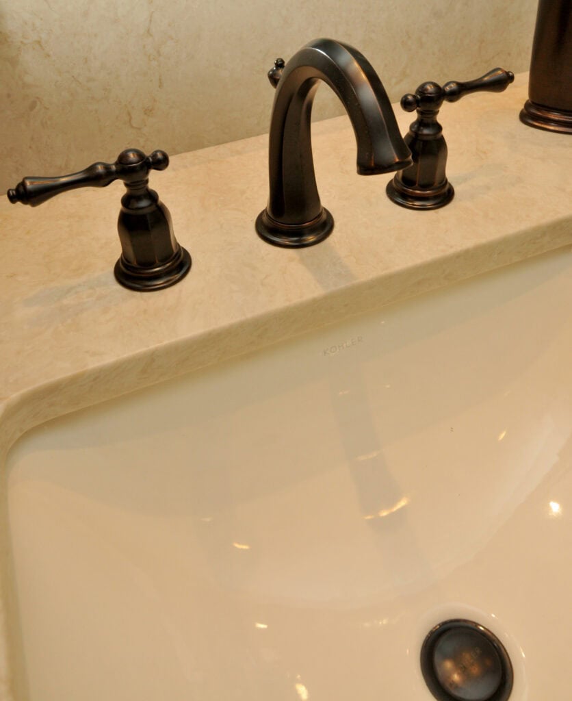 Close-up of a bathroom sink with a marble countertop, bronze faucet, and two handles for hot and cold water. The sink is white and the brand name Kohler is visible inside the basin.