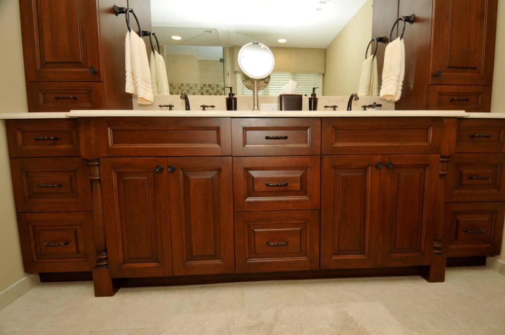 Dark wood double sink bathroom vanity with black hardware, matching cabinets, and drawers. Dual mirrors and towel holders are mounted above the sinks, with beige walls and floor tiles visible.