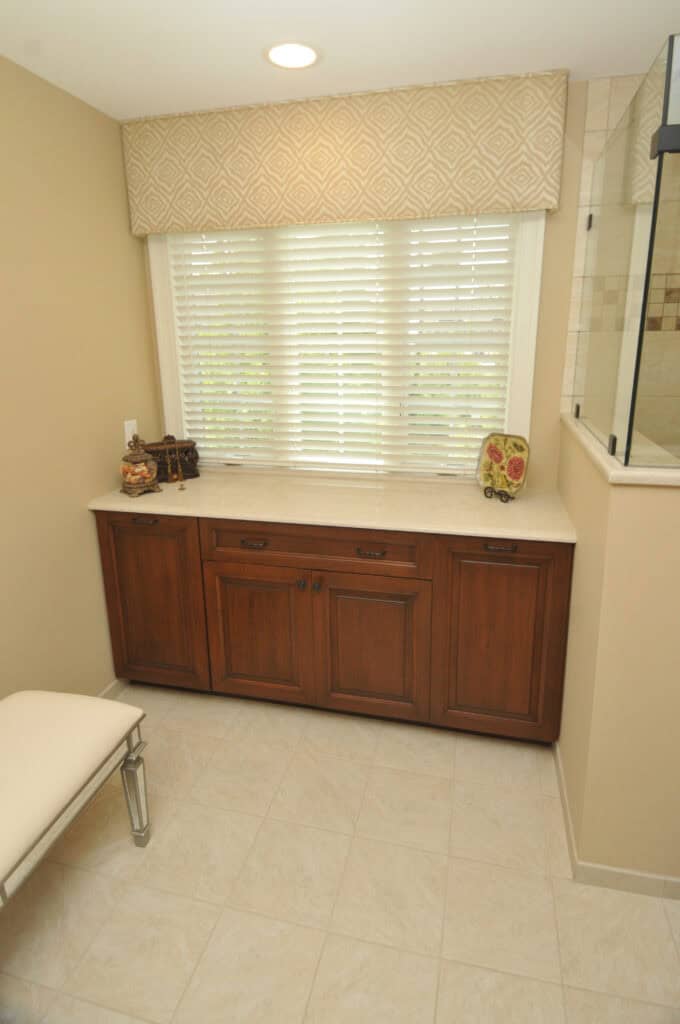 A bathroom features a wooden cabinet with a white countertop beneath a window with white blinds and a patterned valance. Beige walls and tile floor, a cushioned bench, and glass shower enclosure are also visible.