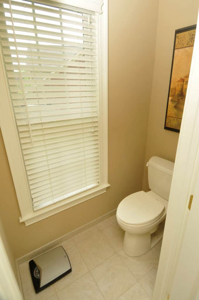 A small bathroom with a white toilet, a window with closed blinds, a wall-mounted painting, light beige walls, and a silver trash can on tiled flooring.