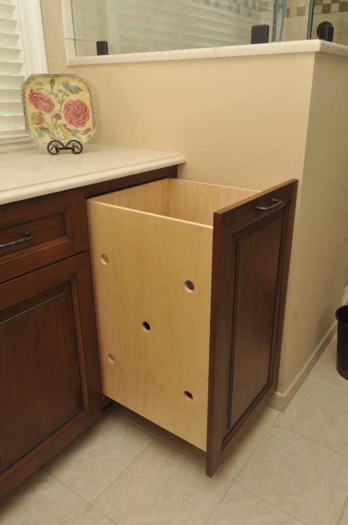 A wooden pull-out laundry hamper with ventilation holes is built into a dark wood bathroom cabinet, partially open beside a countertop with a decorative plate.