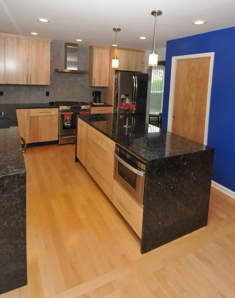 Modern kitchen with light wood cabinets, black granite countertops, stainless steel appliances, hanging pendant lights, light wood floors, and a bright blue accent wall with a wooden door.