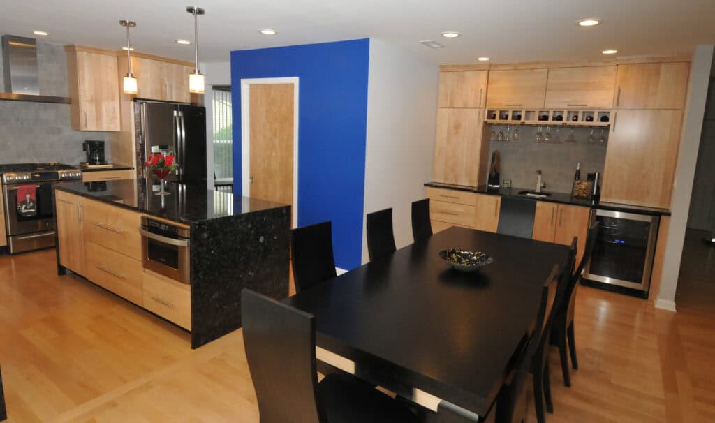 Modern kitchen and dining area with light wood cabinets, black countertops, stainless steel appliances, a black dining table with eight chairs, and a blue accent wall. The room is brightly lit with recessed lighting.