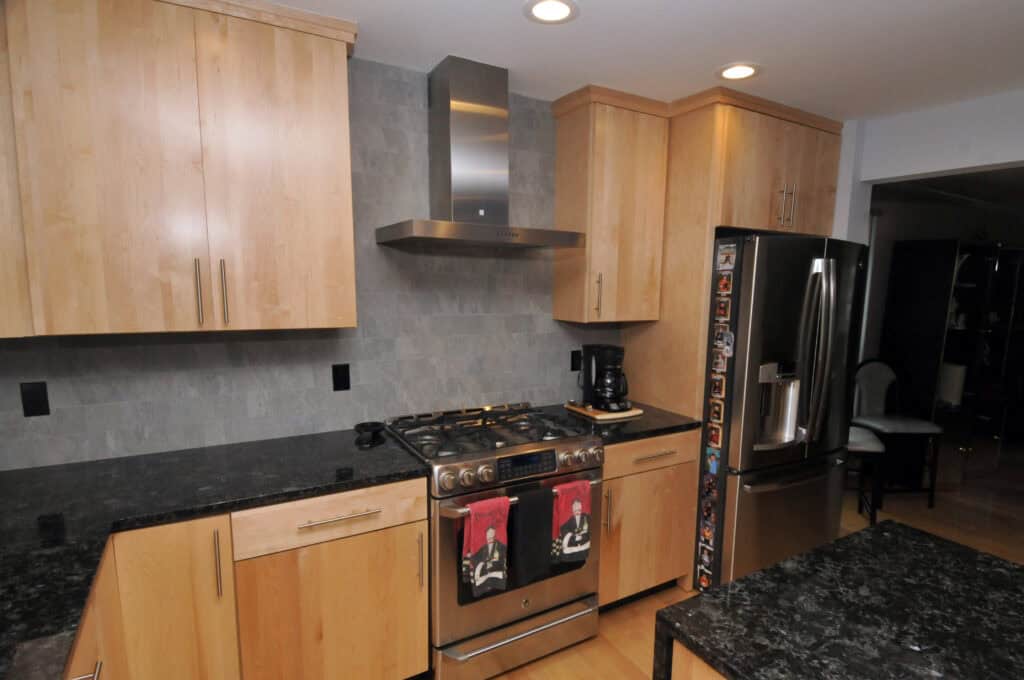 Modern kitchen with light wood cabinets, black granite countertops, stainless steel appliances, a gas stove with an overhead vent, a coffee maker, and a refrigerator with magnets on the side.