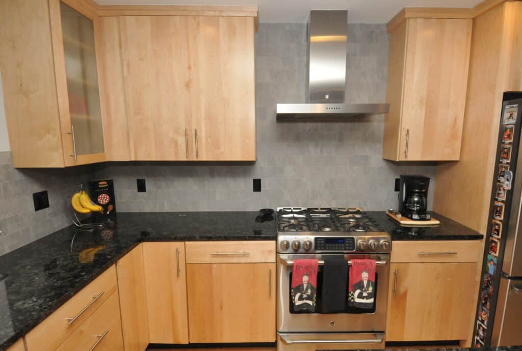 Modern kitchen with light wood cabinets, black granite countertops, stainless steel stove and hood, coffee maker, banana holder, and two decorative towels hanging on the oven door. Refrigerator edge visible on the right.