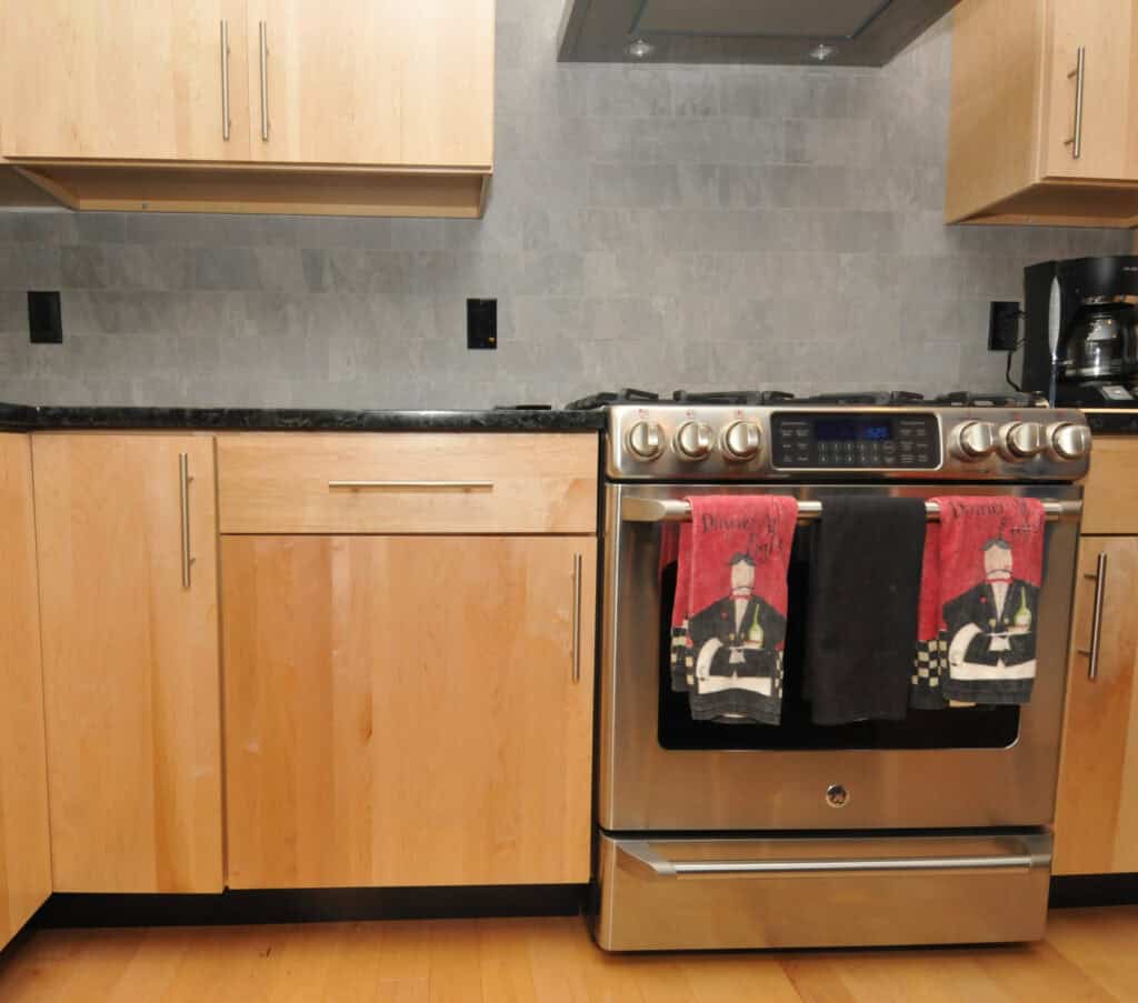 A modern kitchen with light wood cabinets, a stainless steel oven, and two decorative towels hanging on the oven door. The backsplash is gray tile, and a coffee maker sits on the counter to the right.