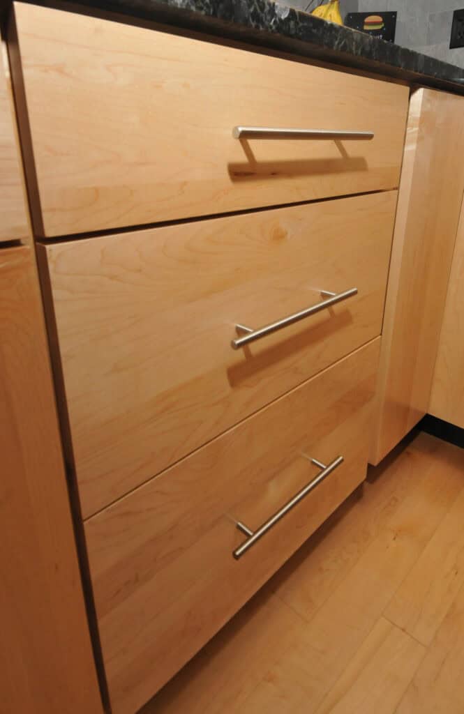 Light wood kitchen drawers with sleek, horizontal metal handles beneath a dark countertop, next to a cabinet. The drawers and cabinet have a modern, minimalist design.