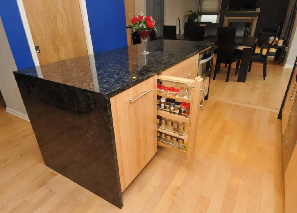 A modern kitchen island with a black granite countertop features a pull-out spice rack drawer, displaying an organized collection of spice jars. A vase with red roses sits on the counter, and a dining area is visible in the background.