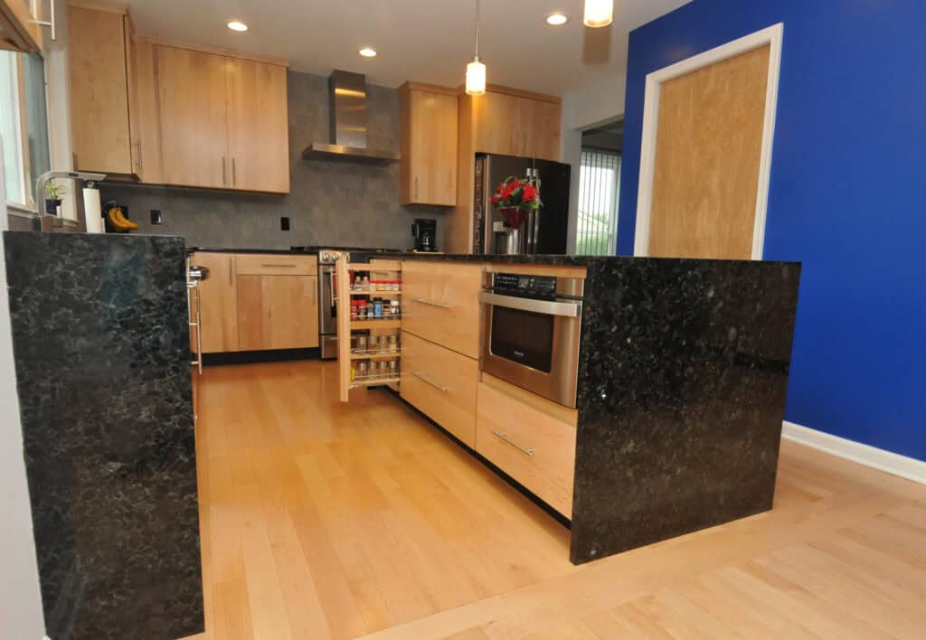 Modern kitchen with light wood cabinets and drawers, black granite countertops, a blue accent wall, a built-in microwave, and a pull-out spice rack. Pendant lights hang from the ceiling.