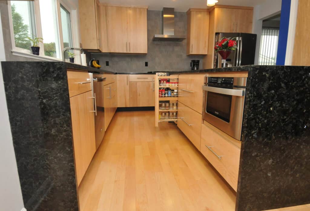 Modern kitchen with light wood cabinets, black granite countertops, light wood flooring, built-in microwave, pull-out spice rack, and small potted plants on the windowsill. A vase of red flowers sits on the counter.