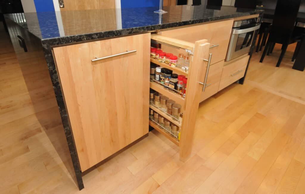 A kitchen with light wood cabinets and a black countertop features a pull-out spice rack filled with spice jars and bottles. The rack is built into the cabinet next to an oven.