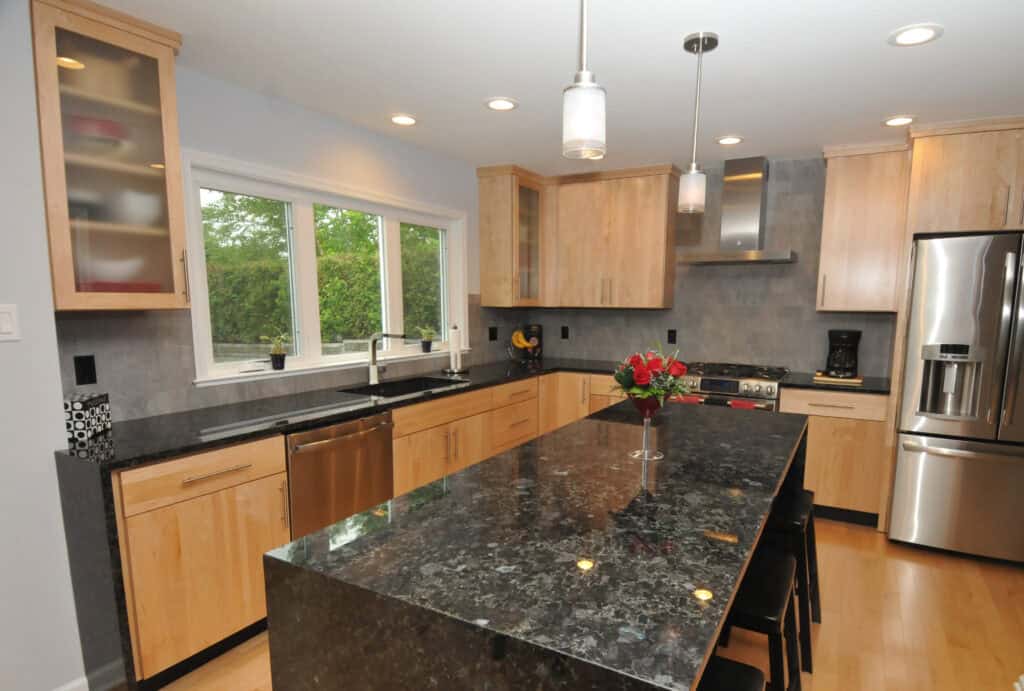 Modern kitchen with light wood cabinets, stainless steel appliances, a large black granite island with chairs, pendant lights, and a window overlooking greenery. A vase with red flowers sits on the island.