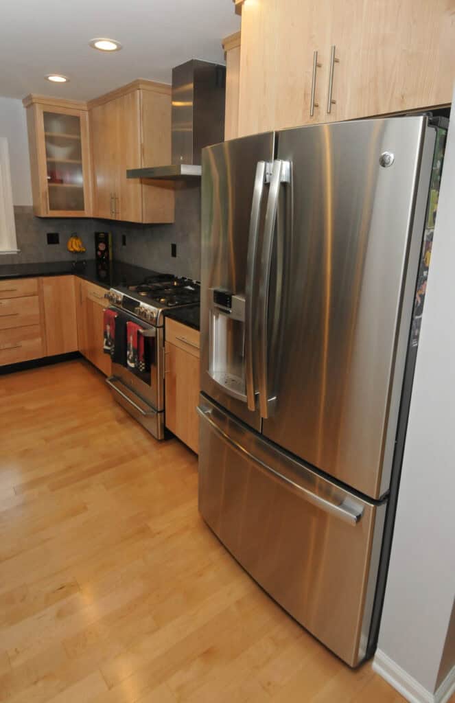 Modern kitchen with light wood cabinets, stainless steel appliances including a large refrigerator with French doors, a stove with an oven, and a microwave. The floor is light wood, and countertops are dark.