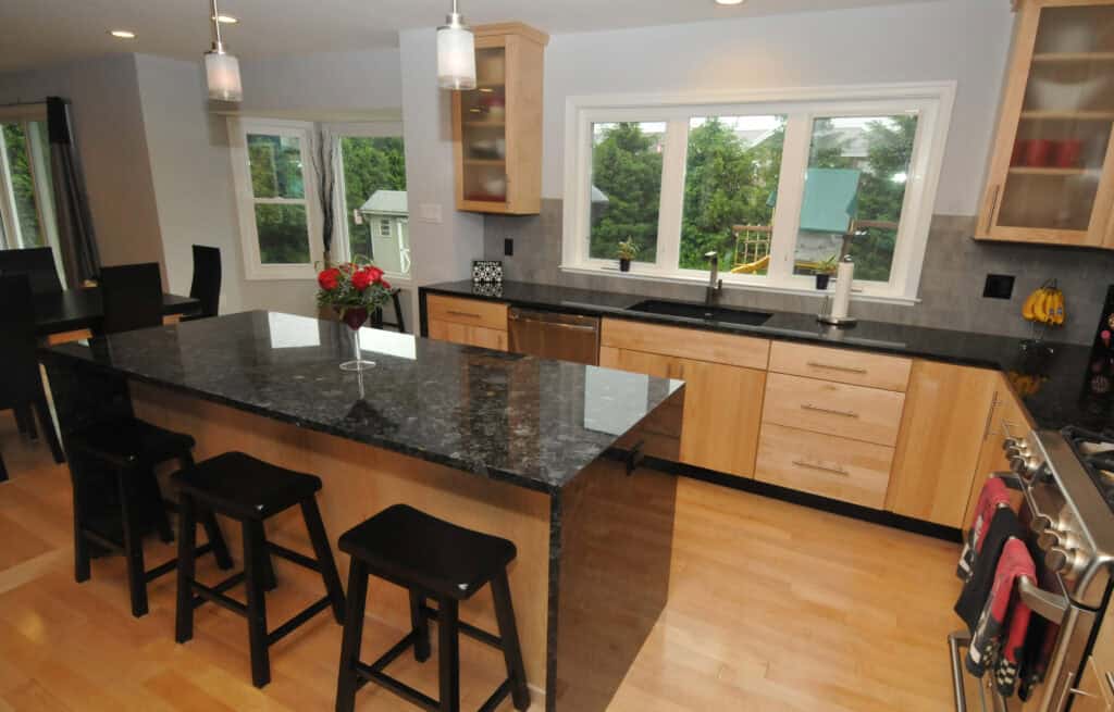 Modern kitchen with light wood cabinets, black granite countertops, an island with three black stools, stainless steel appliances, a vase of flowers, large windows, and light hardwood floors.