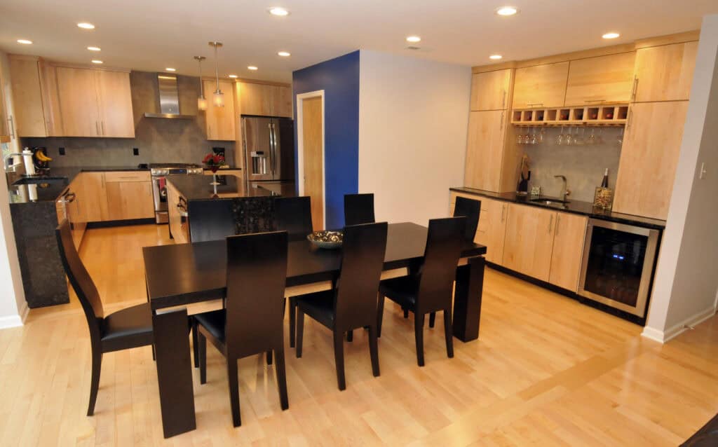 Modern kitchen and dining area with light wood cabinets, a black dining table with eight chairs, stainless steel appliances, built-in wine rack, and recessed lighting on a light wood floor.