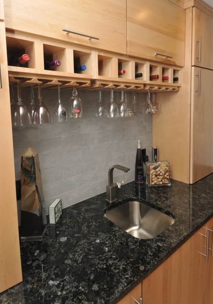 A small home bar with light wood cabinets, a granite countertop, a stainless steel sink, wine bottles stored horizontally above, wine glasses hanging upside down, and assorted bar accessories on the counter.