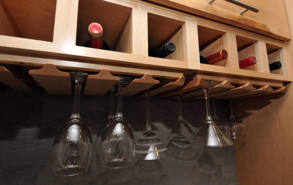 A wooden wine rack mounted under a cabinet holds several bottles of wine horizontally, with clear wine glasses hanging upside down below it.