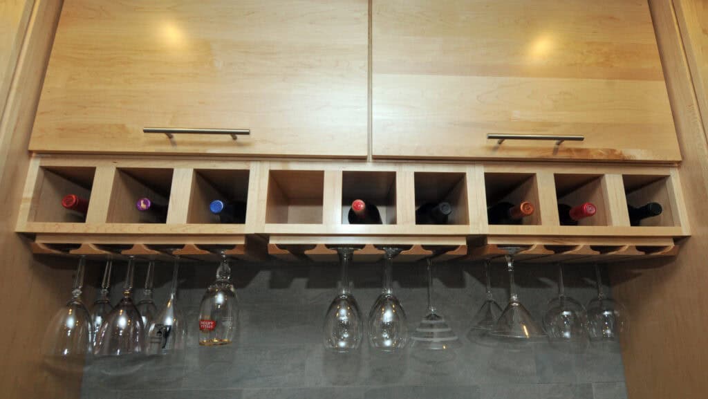 A wooden wine rack holding eight bottles of wine above a row of hanging glassware, including wine and cocktail glasses, mounted on a kitchen wall.
