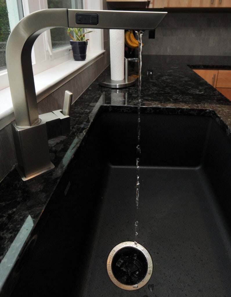 A modern kitchen faucet with a rectangular spout pours water into a black sink set in a dark granite countertop; a roll of paper towels and a potted plant sit by a window in the background.