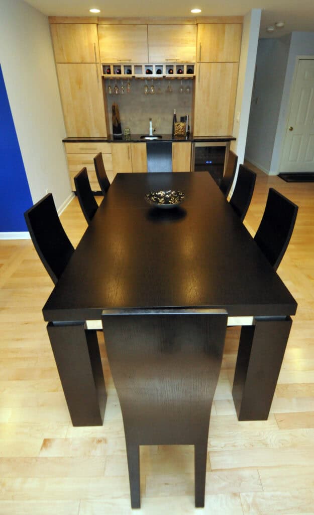 A modern dining room with a long, dark wooden table surrounded by six matching chairs. A decorative bowl sits in the center. Light wood floors and cabinets, with a small bar area and shelves in the background.