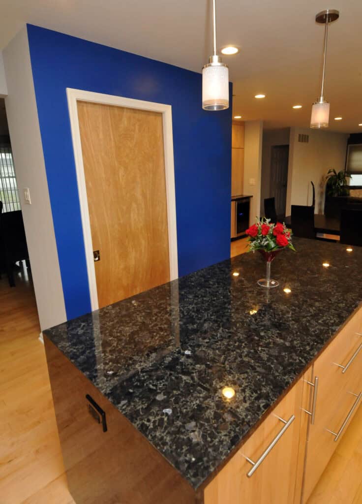 A modern kitchen with a black granite island countertop, light wood cabinets, a vase with red flowers, pendant lights, and a blue accent wall with a wooden door.