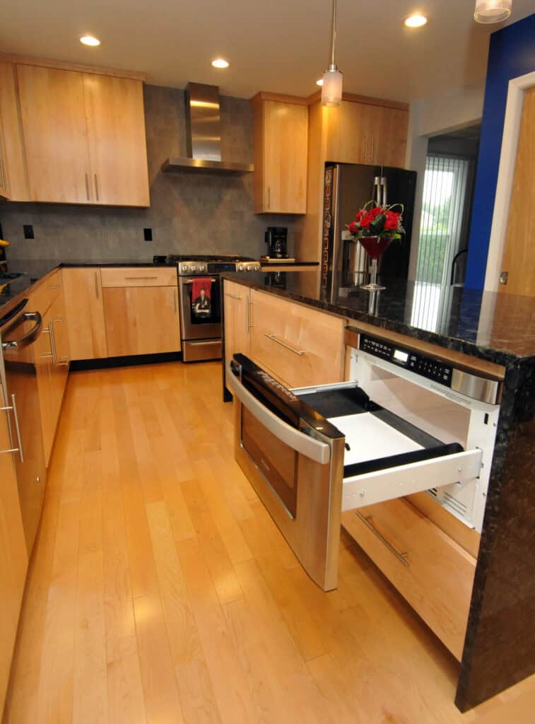 Modern kitchen with light wood cabinets, stainless steel appliances, and black countertops. A drawer-style microwave is open under the counter, and a vase with red flowers sits on the island.