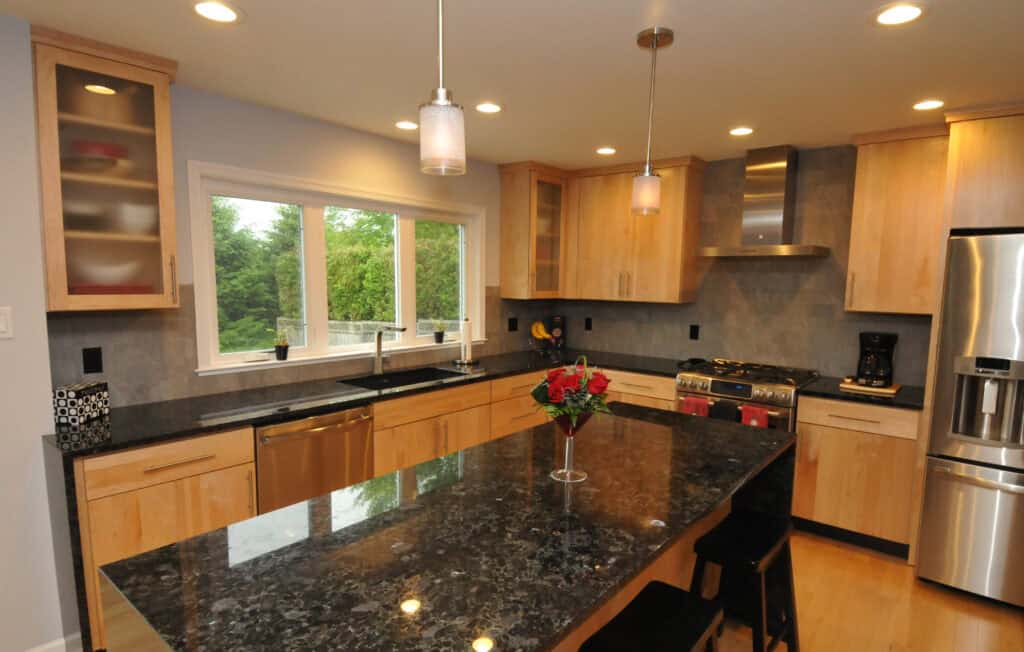 Modern kitchen with light wood cabinets, stainless steel appliances, a dark granite island with two stools, hanging pendant lights, and a vase with red flowers. Large window provides natural light and a view of greenery.