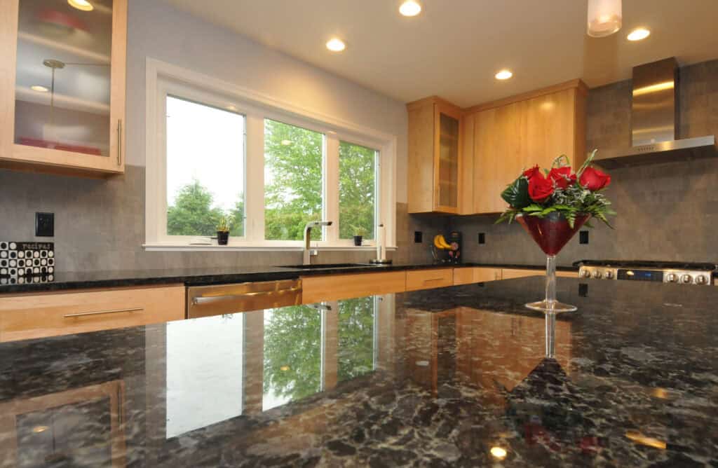 Modern kitchen with black granite countertops, light wood cabinets, large window over the sink, stainless steel appliances, recessed lighting, and a vase with red roses on the counter.