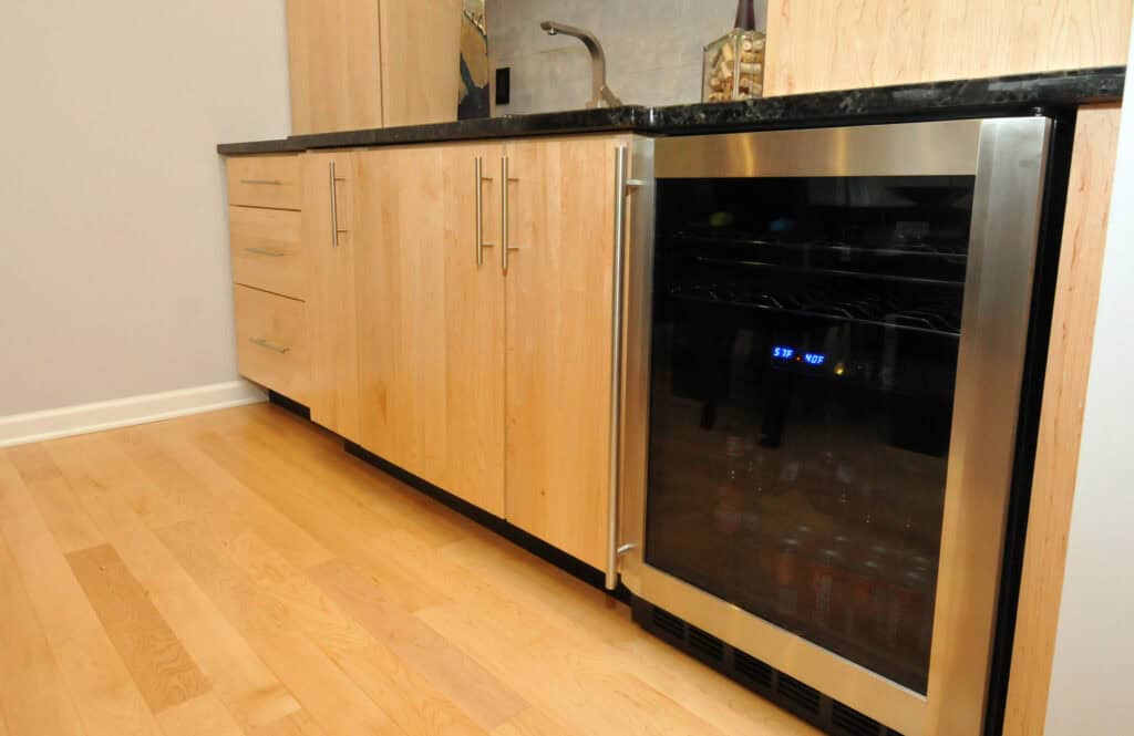A modern kitchen with light wooden cabinets, black countertop, and a stainless steel mini fridge with a glass door displaying drinks inside, set on a light hardwood floor.