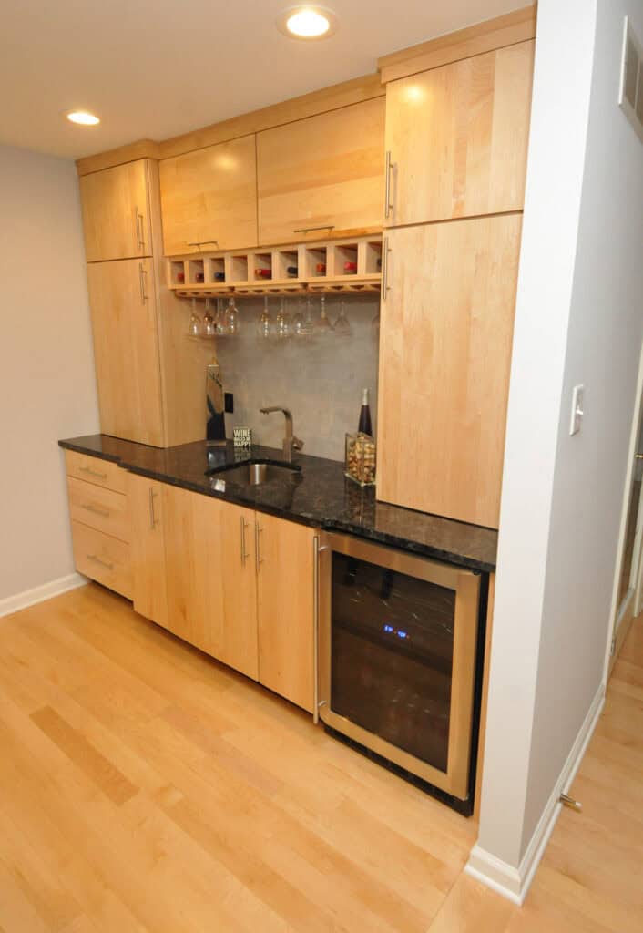 A modern kitchenette with light wood cabinets, a black countertop, a small sink, a built-in wine rack with hanging glasses, and a glass-front mini fridge. The floor and walls are light-colored.