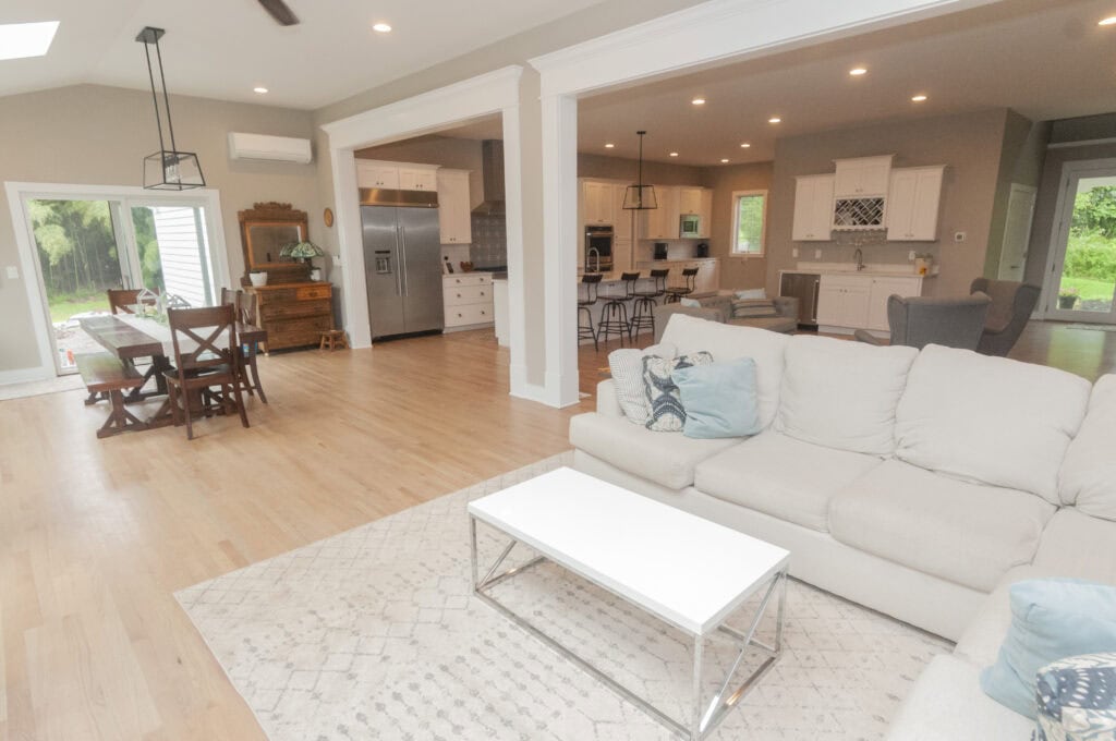 Open-concept living space with a light-colored sofa, coffee table, and rug in the foreground; a dining table and modern kitchen with bar stools and white cabinets are in the background. Large windows let in natural light.