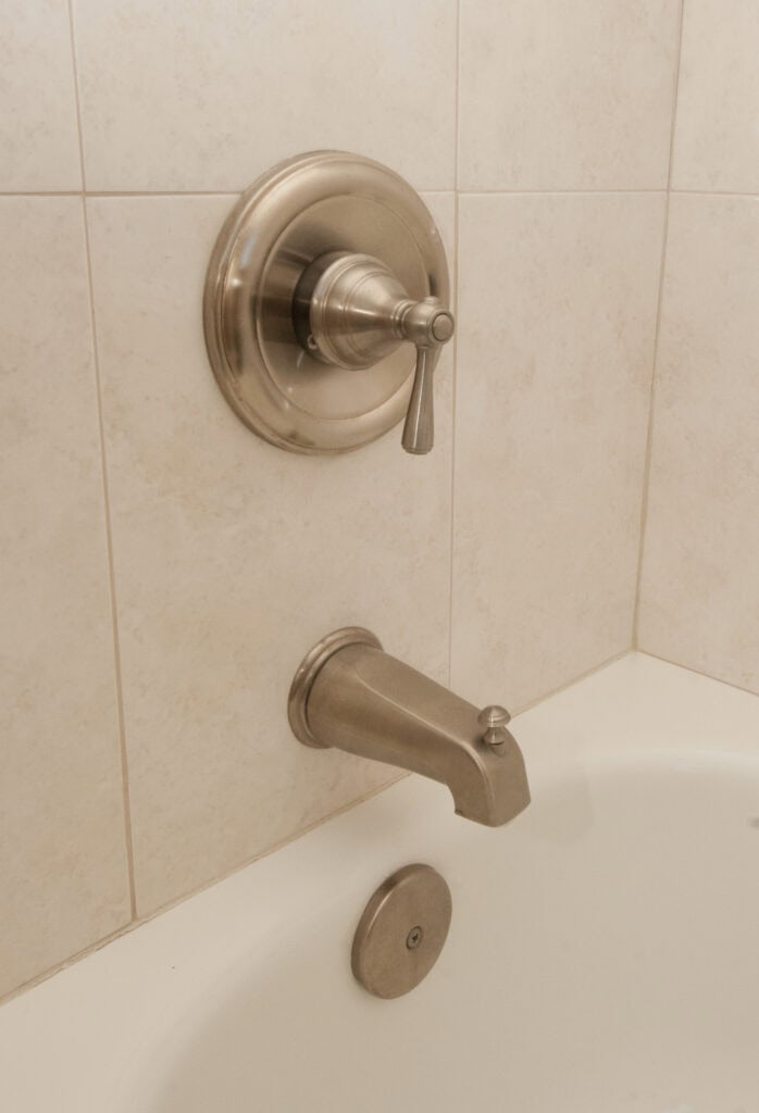 A close-up of a bathtub faucet and single-handle shower control mounted on a tiled bathroom wall, with part of the white bathtub and drain visible at the bottom.