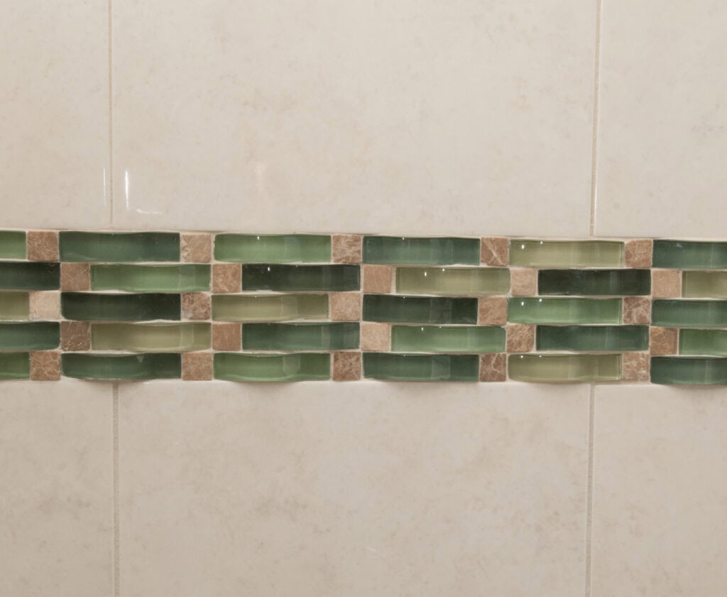 A bathroom wall with beige tiles, featuring a horizontal decorative strip of green and brown rectangular mosaic tiles.
