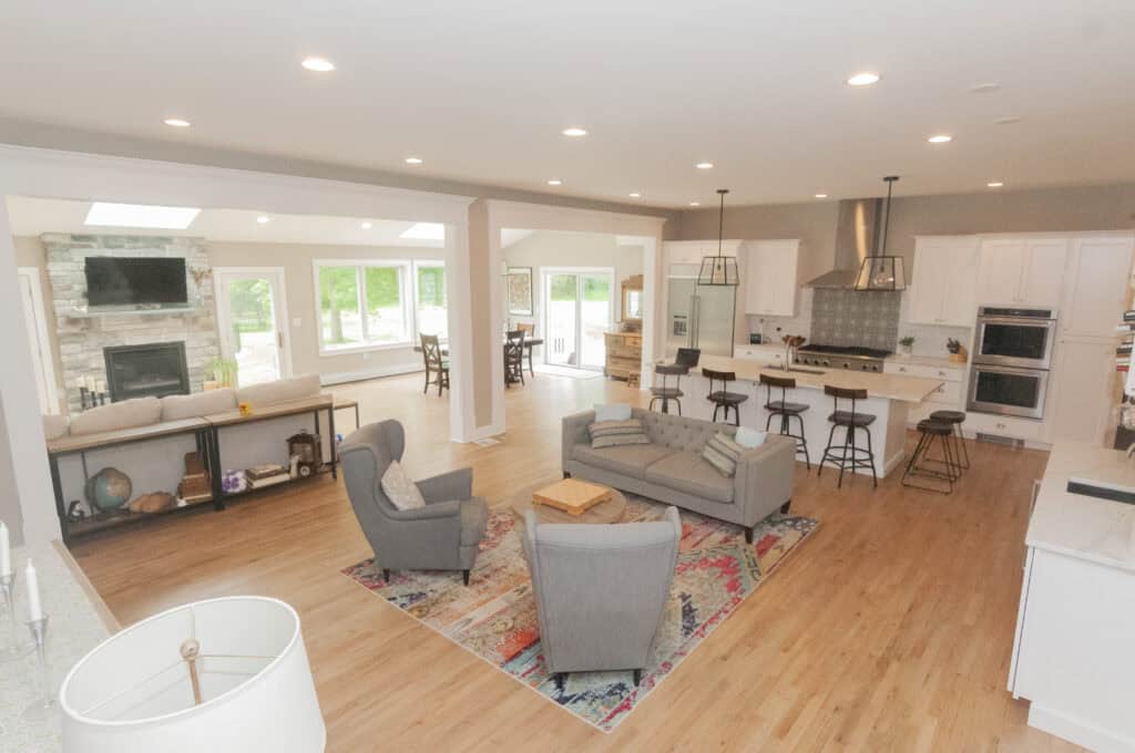 Open-concept living area with a kitchen, island seating, gray furniture on a colorful rug, dining table, large windows, and a stone fireplace. Light wood flooring and recessed lighting throughout the space.