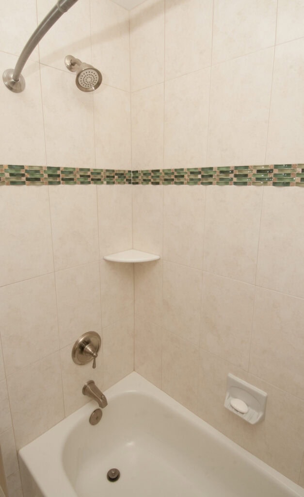 A white tiled bathtub and shower with a silver showerhead, faucet, a small corner shelf, green tile accent strip, and a soap holder with a bar of soap.