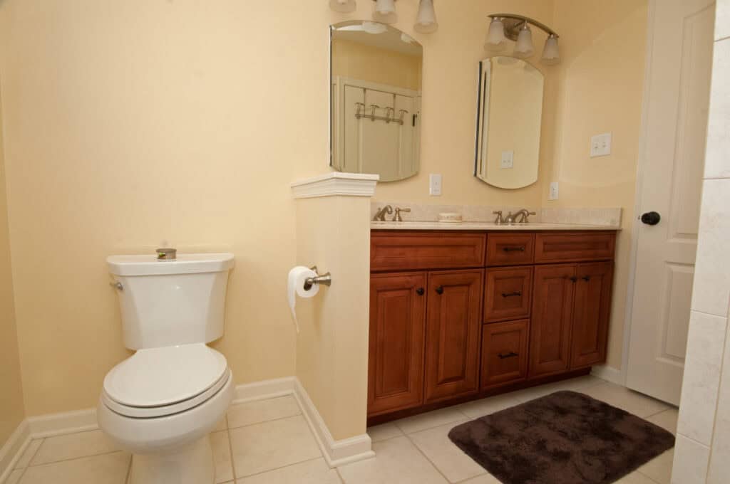 A bathroom with a white toilet, toilet paper holder, wooden vanity with two sinks, two mirrors, light fixtures above, and a brown rug on the tiled floor against beige walls.