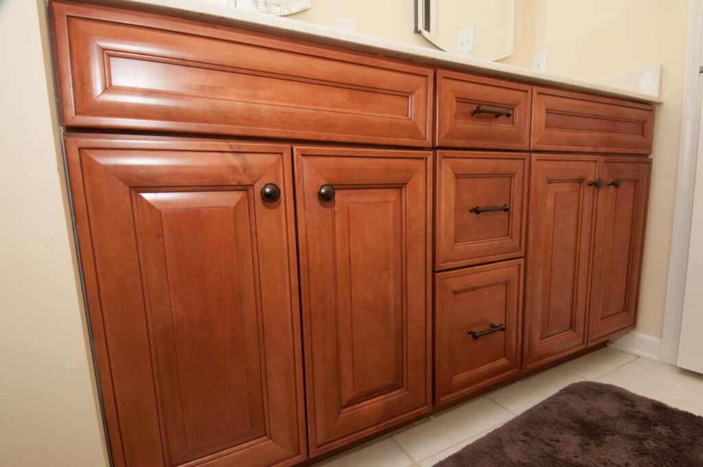 Wooden bathroom vanity with a light-colored countertop, featuring three drawers and four cabinet doors with dark metal handles and knobs, set against a tiled floor and beige wall.