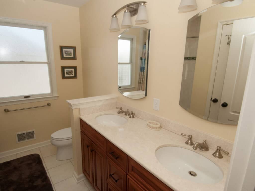A bathroom with a double sink vanity, two mirrors, brushed nickel fixtures, a toilet near a frosted window, two small framed pictures, and a dark rug on a light tile floor.