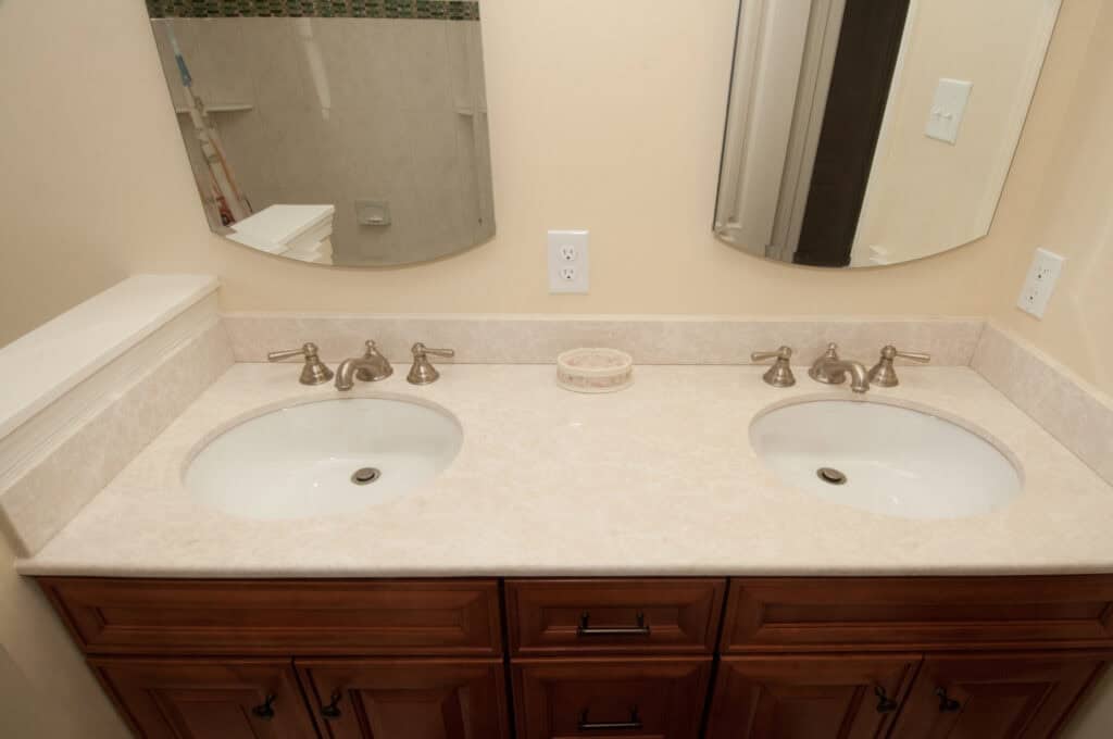 Double bathroom vanity with two white sinks, beige marble countertop, bronze faucets, wooden cabinets, two mirrors, and a small decorative dish between the sinks. Cream-colored walls and partial view of a shower in the background.
