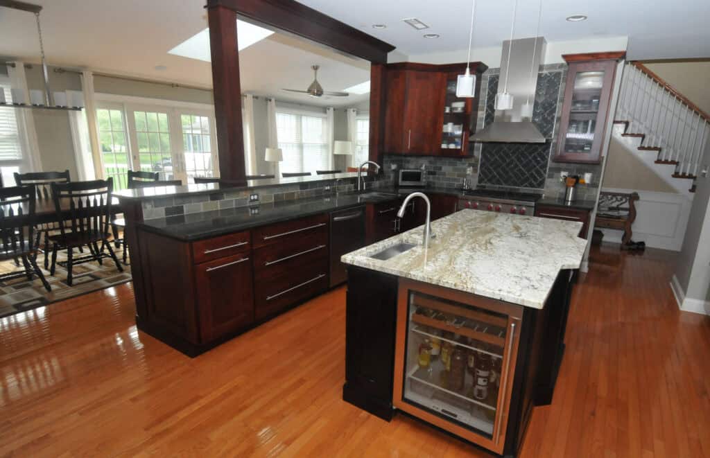 Modern kitchen with dark wood cabinets, a granite island countertop with a sink, built-in wine fridge, stainless steel appliances, tile backsplash, and hardwood floors; dining area and staircase visible in the background.
