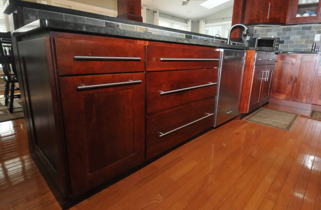 A modern kitchen features dark wood cabinets and drawers with sleek silver handles, a black countertop, stainless steel appliances, and shiny wooden flooring.