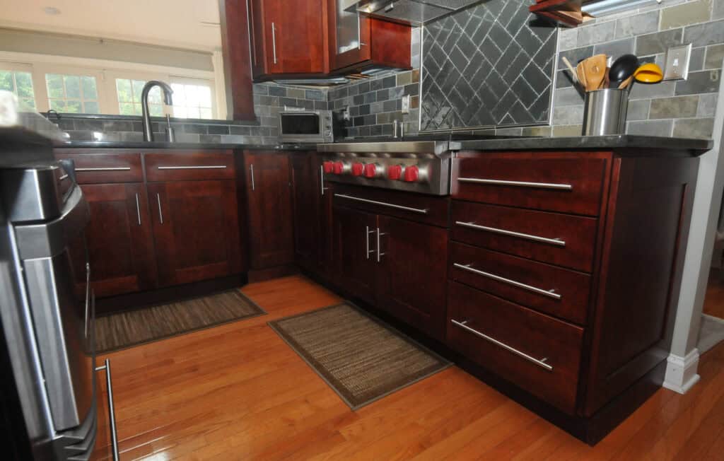 Modern kitchen with dark wood cabinets, stainless steel handles, black countertops, and a stove with red knobs. The backsplash features dark tiles, and there are small rugs on the shiny wooden floor.