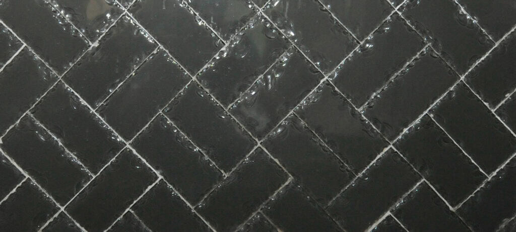 Glossy black rectangular tiles arranged in a diagonal herringbone pattern, with white grout lines highlighting the geometric design. The surface appears wet or reflective, giving it a shiny look.