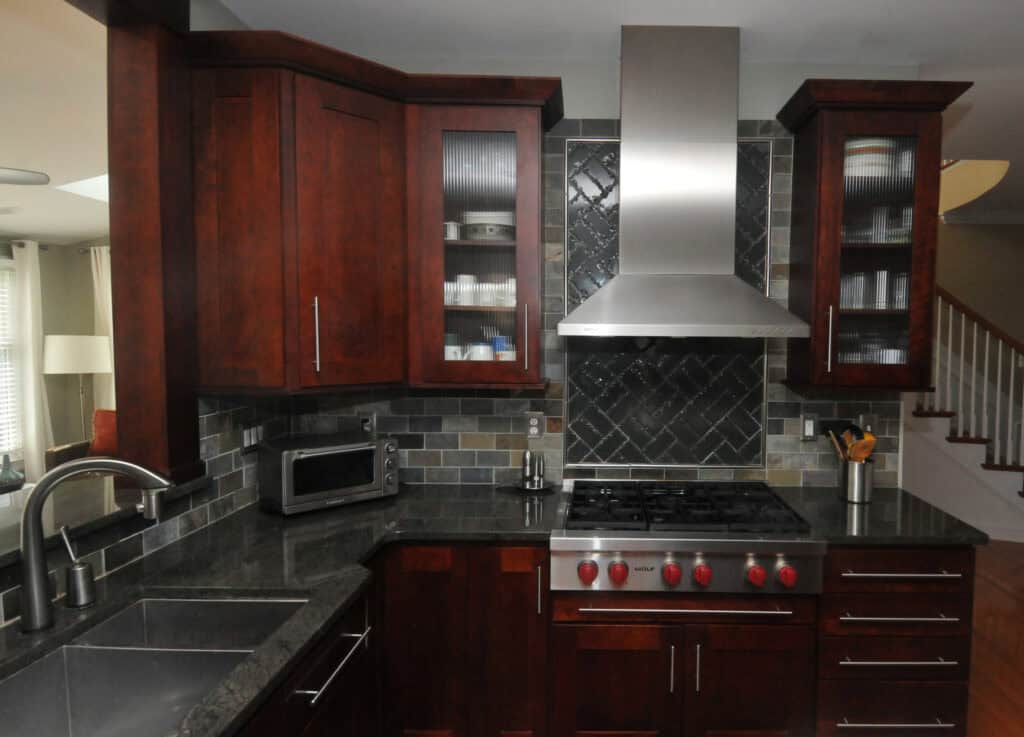 Modern kitchen with dark wood cabinets, stainless steel appliances, glass cabinet doors displaying dishes, a tile backsplash, and a gas stove with a large metal hood. Sink and faucet are visible on a black countertop.