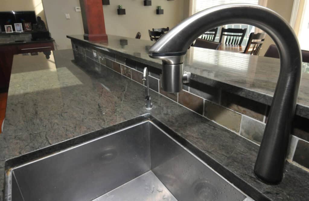 Close-up of a modern kitchen sink with a stainless steel faucet and soap dispenser set in a dark stone countertop. In the background, dining chairs and a window with blinds are visible.