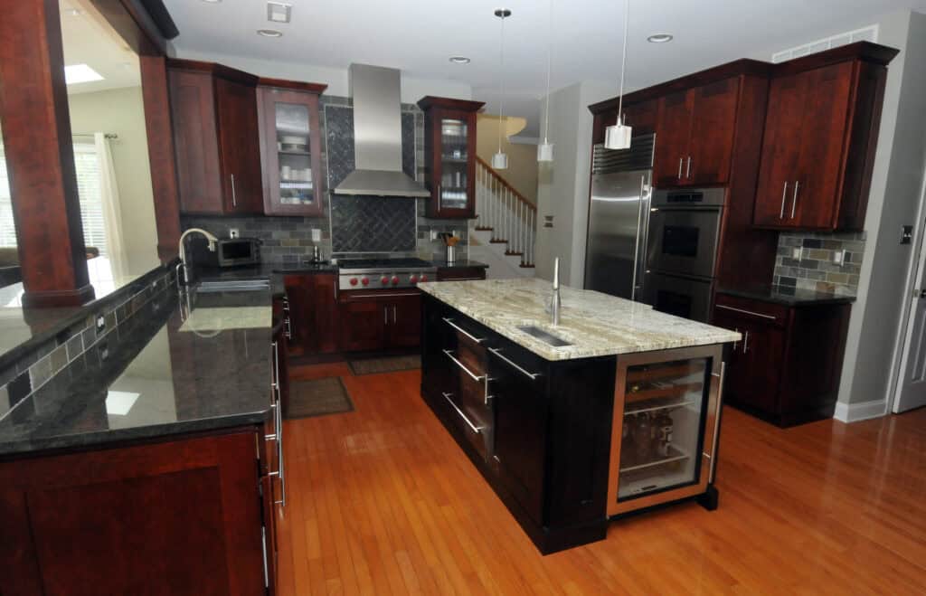 Modern kitchen with dark wood cabinets, stainless steel appliances, a large island with a sink and mini fridge, granite countertops, hardwood floor, tiled backsplash, and pendant lighting.
