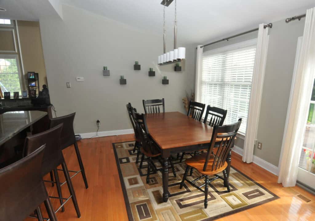 A dining room with a wooden table, six black chairs, a geometric rug, and a modern rectangular chandelier. There are small wall planters on a light gray wall and large windows with white blinds and curtains.