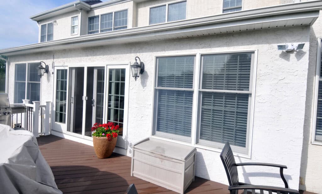 A sunny patio with wooden decking, a covered grill, black outdoor chair, storage box, potted red flowers, and double glass doors leading into a white house with large windows and white blinds.