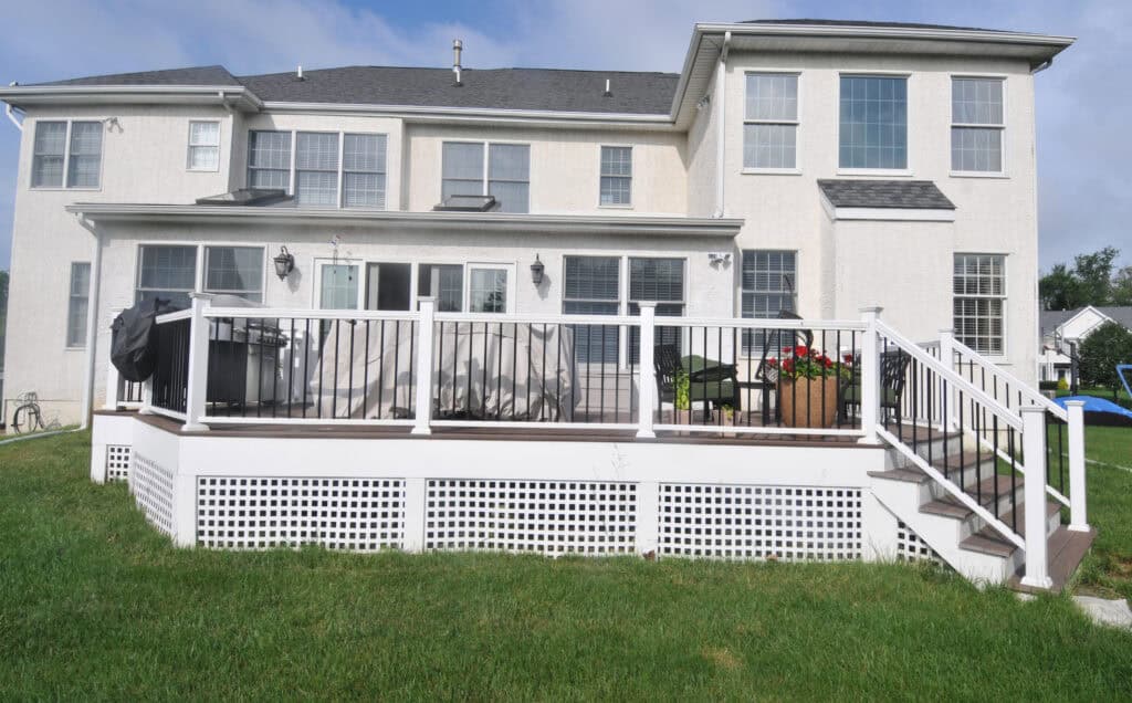 A large white two-story house with many windows and a spacious backyard. The house features a raised wooden deck with white railing, outdoor furniture, a grill, and potted plants on a green lawn.