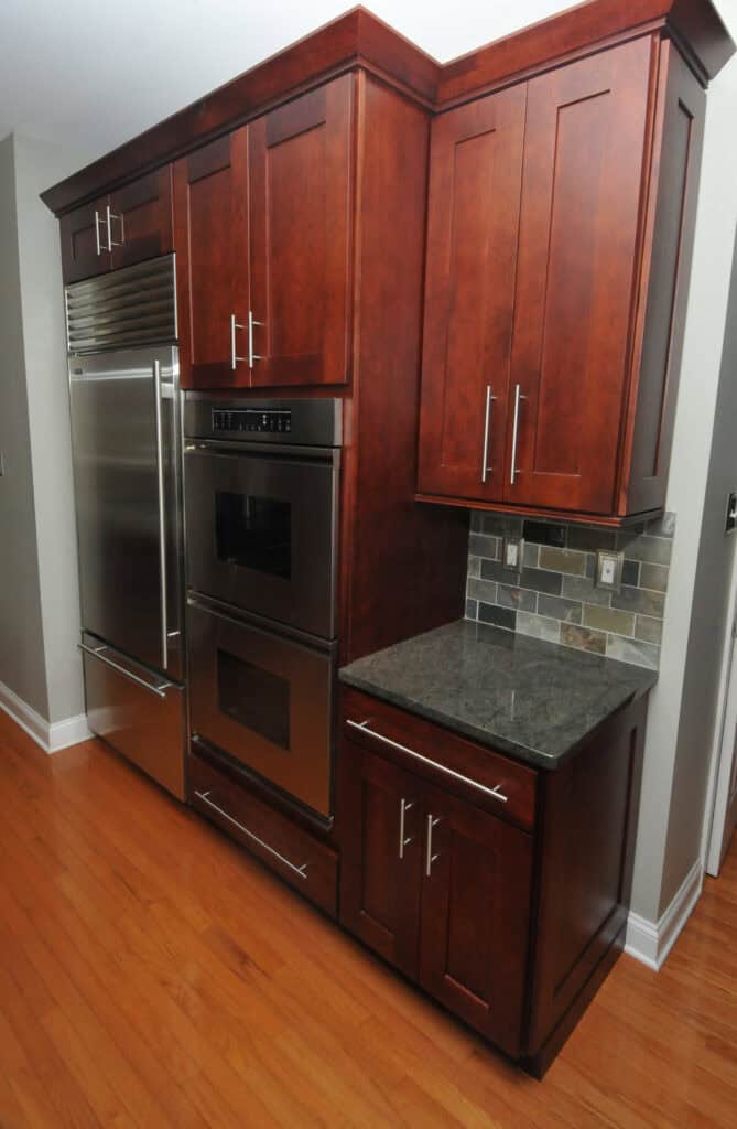 Kitchen with dark wood cabinets, stainless steel built-in double ovens, a large refrigerator, gray stone countertop, and tiled backsplash, all set against a light-colored wall and wooden floor.